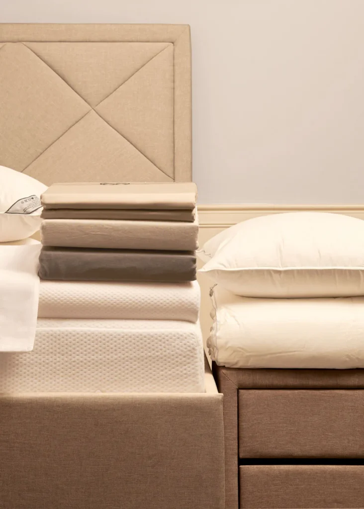 Gestapeld bedlinnen in bruine en beige tinten met witte kussens op een boxspring met gestoffeerd hoofdbord
