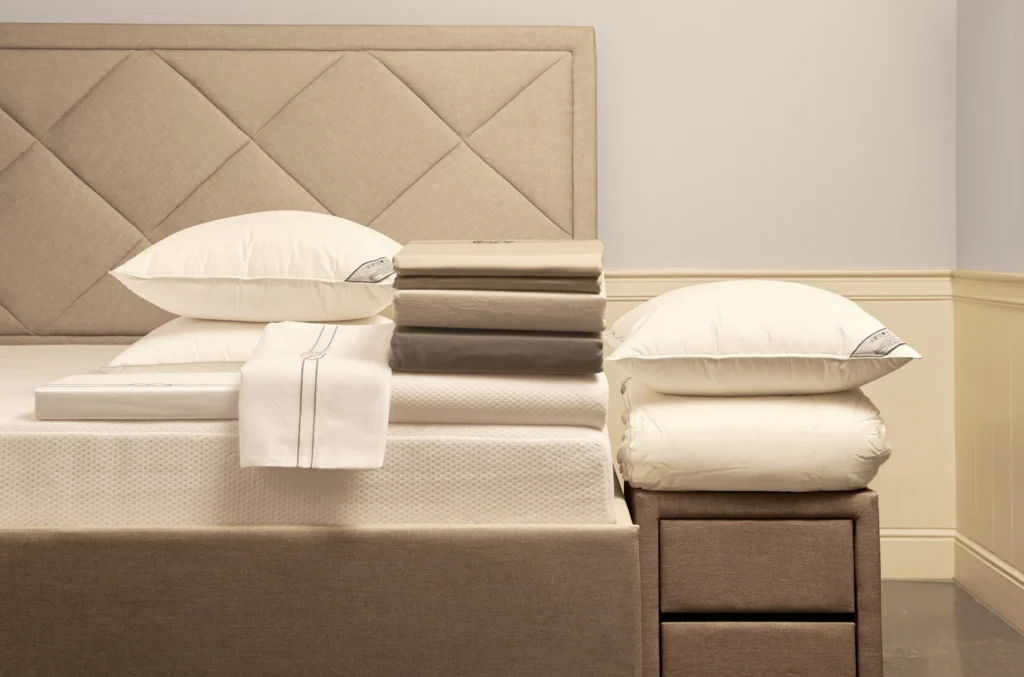 Boxspring met gestoffeerd hoofdbord, gestapeld bedlinnen in zachte tinten en witte hoofdkussens