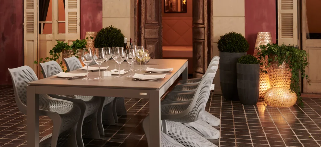 Brede kijk op een gedekte buitentafel met witte stoelen voor een mediterrane gevel met luiken, planten en lantaarns