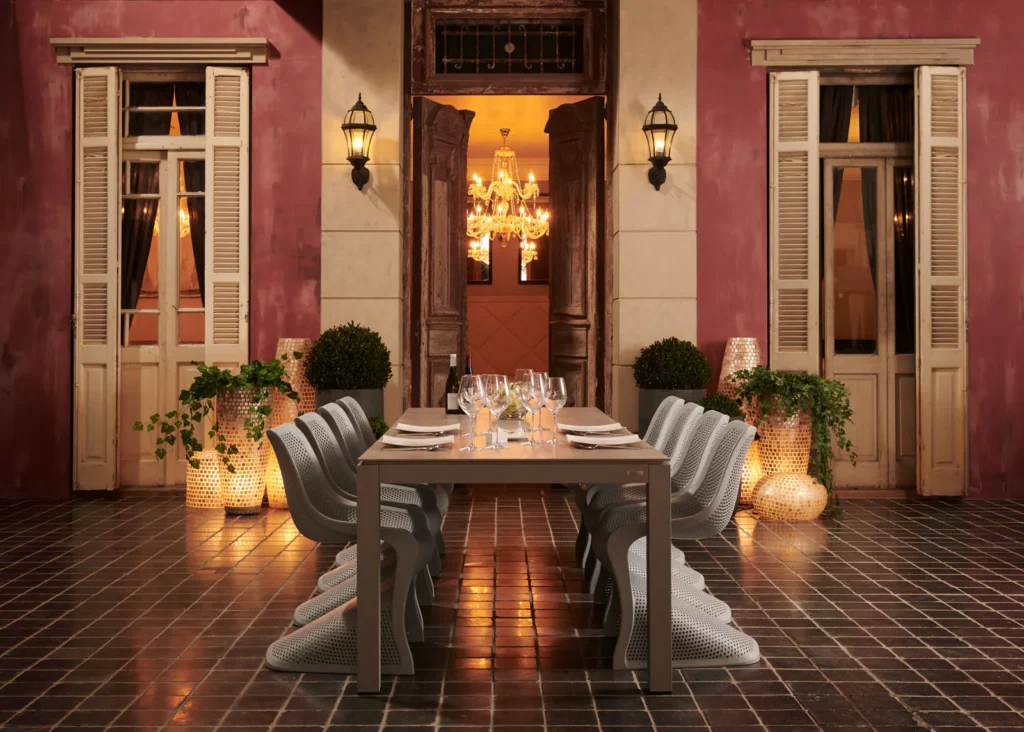 Gedekte buitentafel met witte stoelen op een terras voor een roze mediterrane gevel, met lantaarns en kaarslicht bij avond