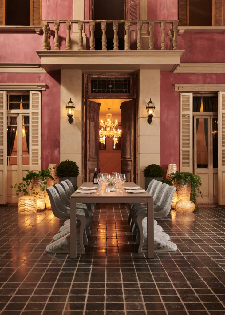 Lange gedekte buitentafel met witte stoelen voor een roze gevel met balkon, lantaarns en kaarsen bij avond