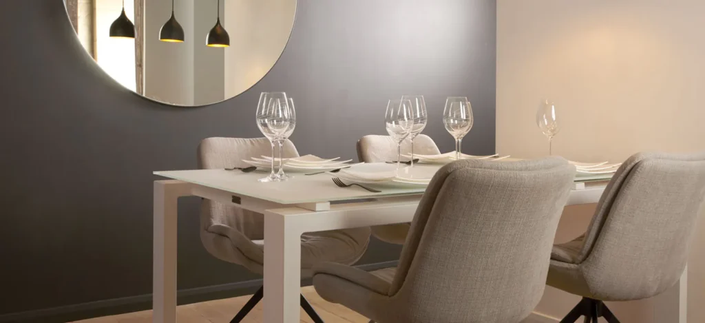 Eetkamer met een witte tafel, gedekte glazen, beige stoffen draaistoelen, zwarte hanglampen en een ronde spiegel