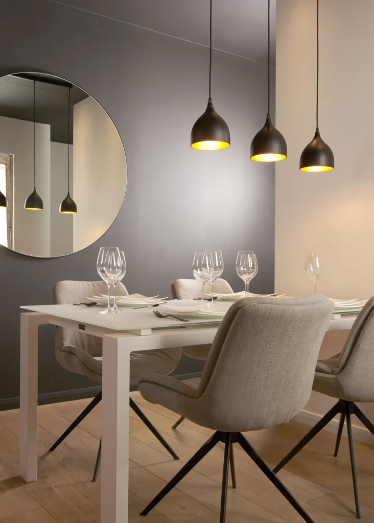 Eetkamer met witte tafel, beige stoffen stoelen op zwarte poten, drie zwarte hanglampen en een grote ronde spiegel