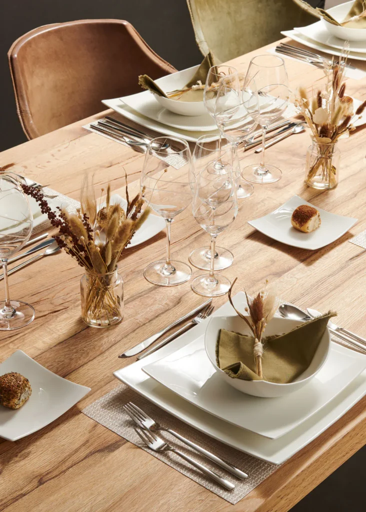 Bovenaanzicht van een gedekte eiken tafel met wit servies, gevouwen servetten, kristallen glazen en droogbloemen
