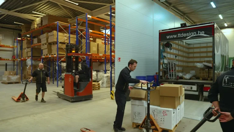 Medewerkers zetten dozen klaar bij een vrachtwagen van T&T Design, met een heftruck tussen de palletrekken in het magazijn