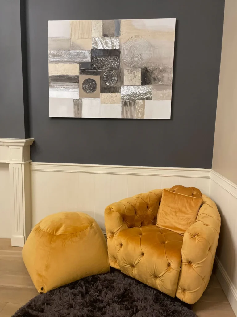 Okergele fluwelen capitonfauteuil met bijpassende poef op een donker hoogpolig tapijt, onder een abstract schilderij