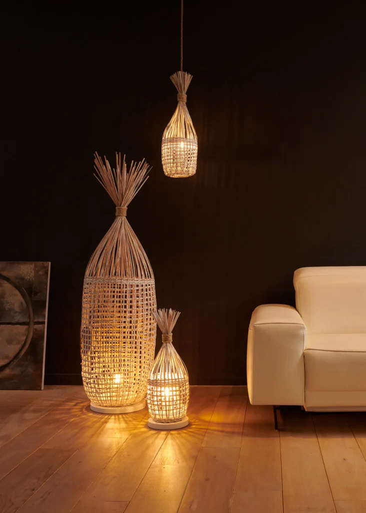 Twee rieten vloerlampen en een rieten hanglamp met warm licht naast een lichte lederen zetel op een houten vloer
