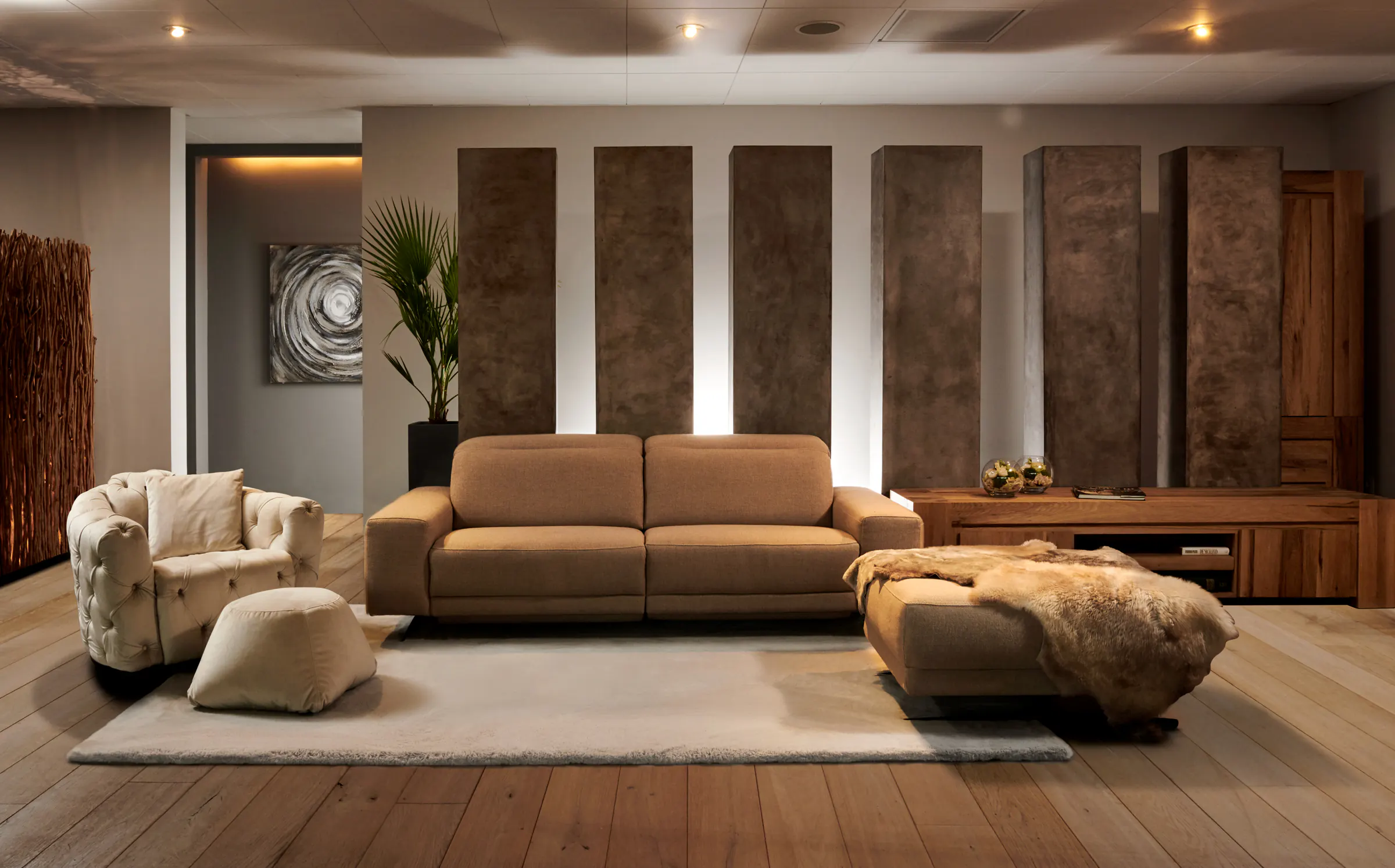 Salon met cognackleurige driezit, capitonfauteuil, poef met vacht en houten dressoir voor bronskleurige wandpanelen