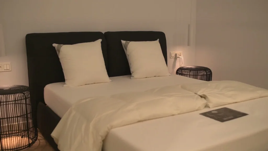 Bed met zwart gestoffeerd hoofdbord, wit bedlinnen en witte kussens naast rieten lampjes in een lichte slaapkamer