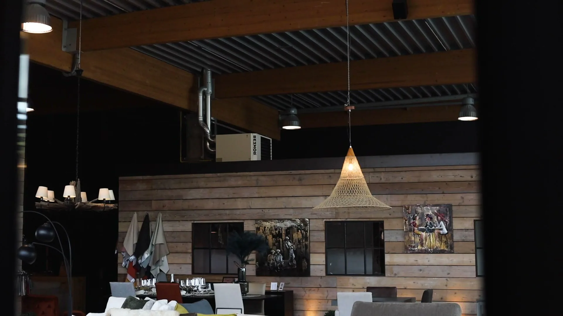 Showroom met hoge zwarte balken, een houten wand met kunst, hanglampen en gedekte tafels