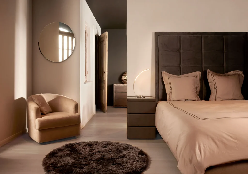 Slaapkamer met donker gestoffeerd hoofdbord, zachtroze bedlinnen, kleine fauteuil en ronde spiegel aan de muur