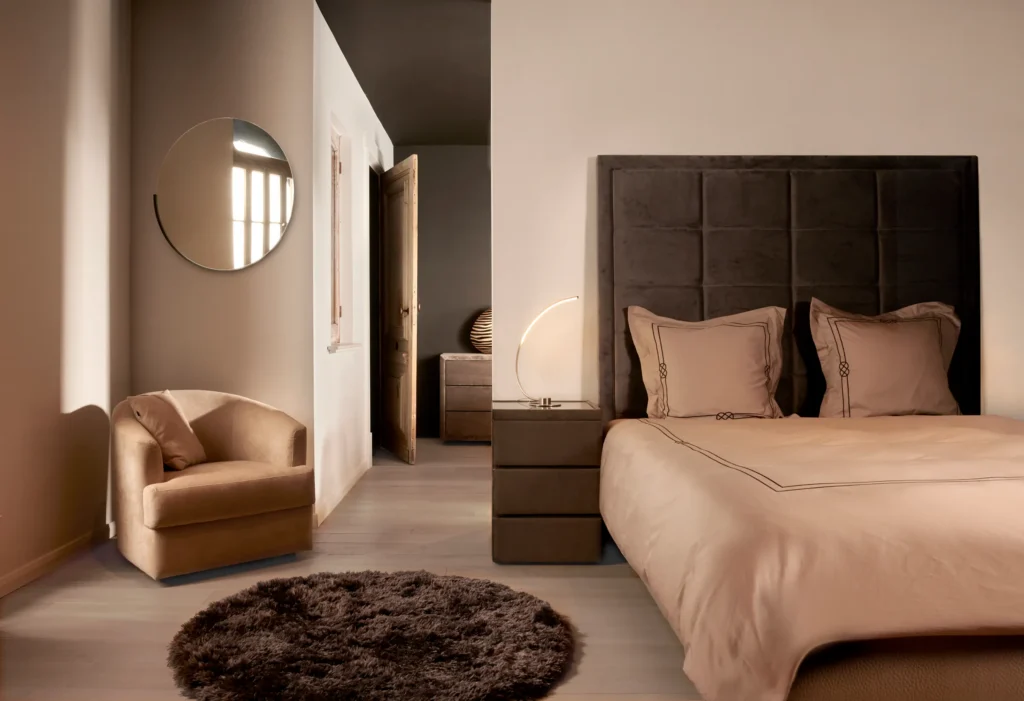 Slaapkamer met hoog gecapitonneerd hoofdbord in bruine stof, beige bedlinnen, fauteuil, nachtkastjes en ronde spiegel