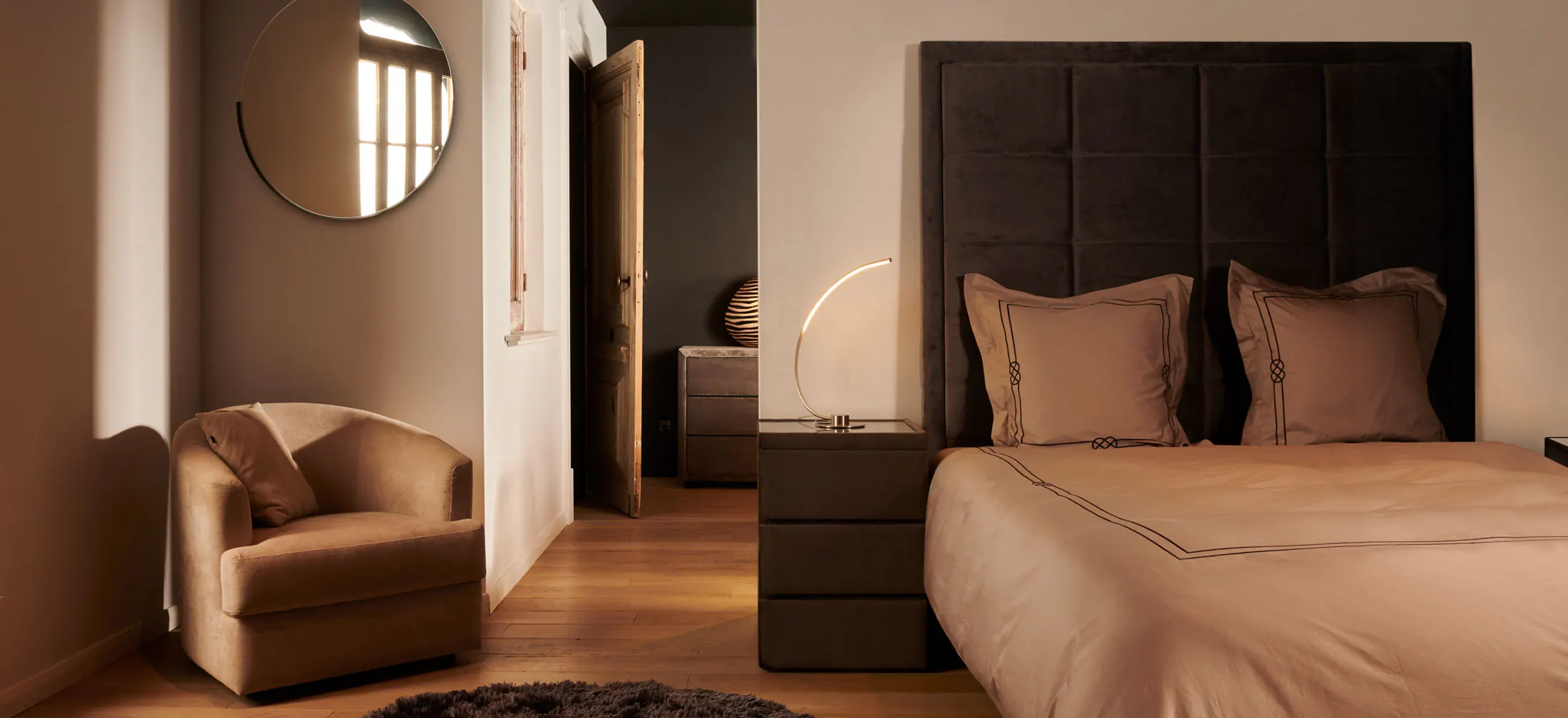 Brede kijk op een slaapkamer met donker gestoffeerd hoofdbord, beige bed, fauteuil en ronde spiegel in warm licht