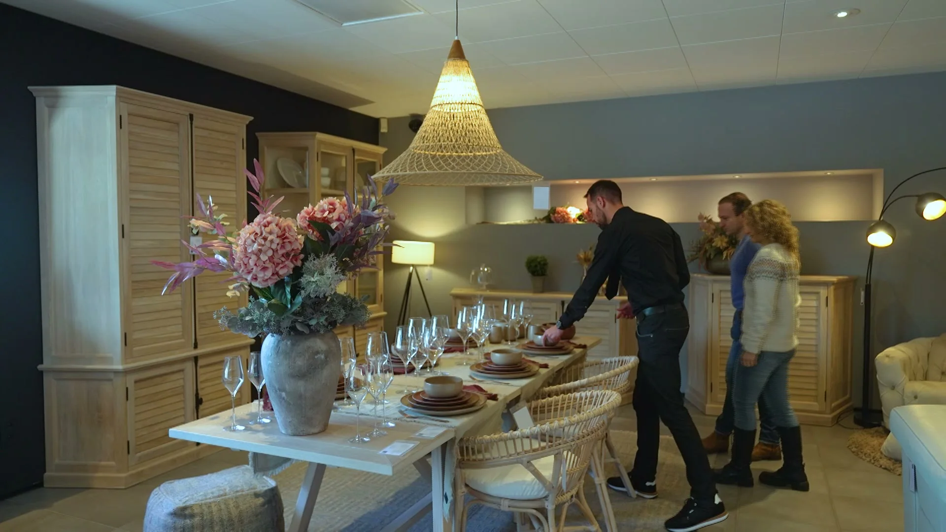Adviseur van T&T Design dekt een lange tafel terwijl een koppel meekijkt in de showroom, onder een rieten hanglamp