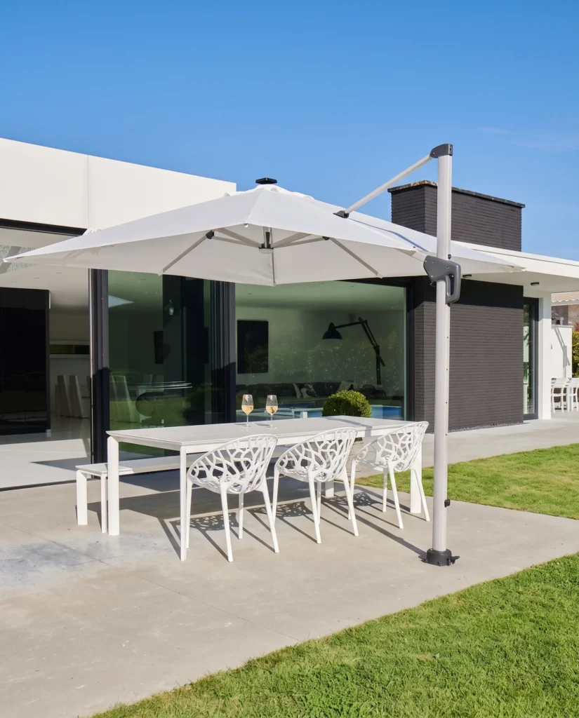 Witte zweefparasol boven een lange witte tuintafel met bank en opengewerkte stoelen op een zonnig terras bij een moderne woning